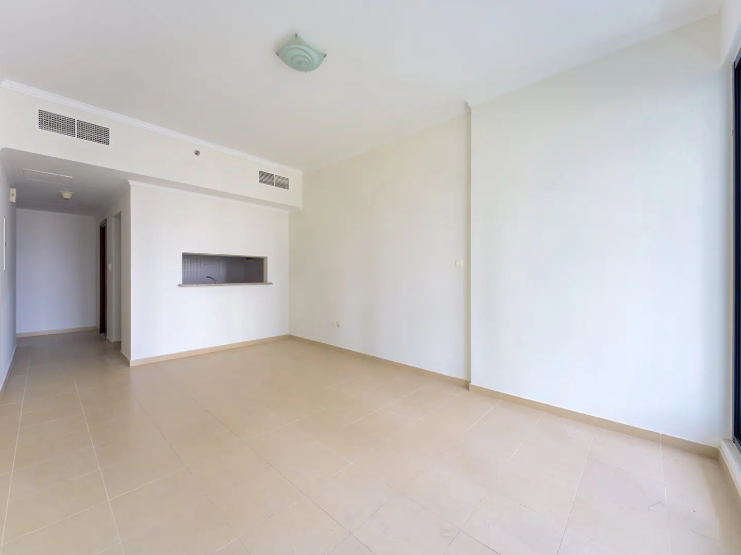 Empty living room with white walls, beige tile floors, and a kitchen pass-through window.