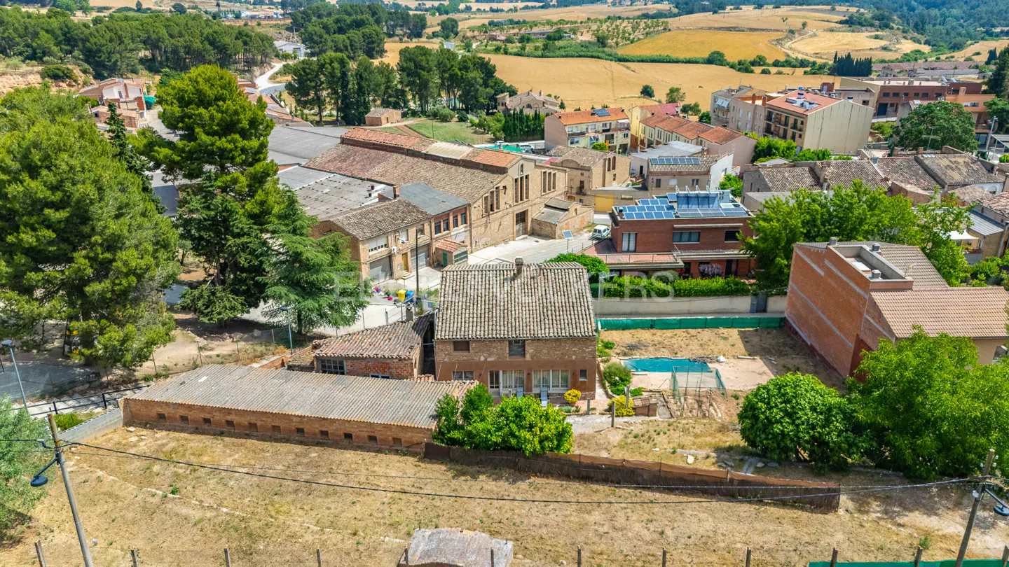 Aerial view of a red brick house with a pool in a rural town, surrounded by trees and fields.