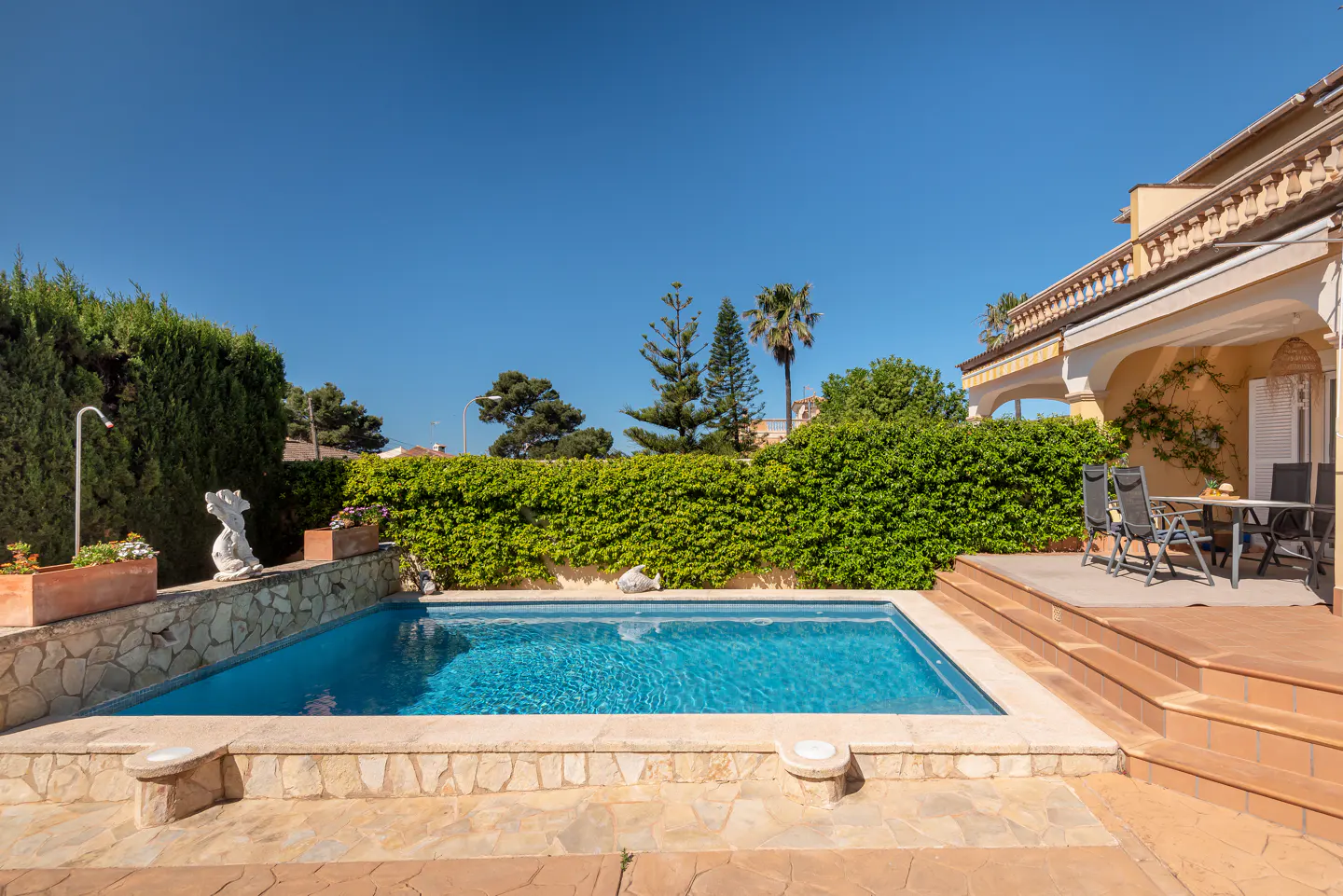 A rectangular swimming pool with blue water sits on a stone patio next to a house with a covered terrace and green hedge.