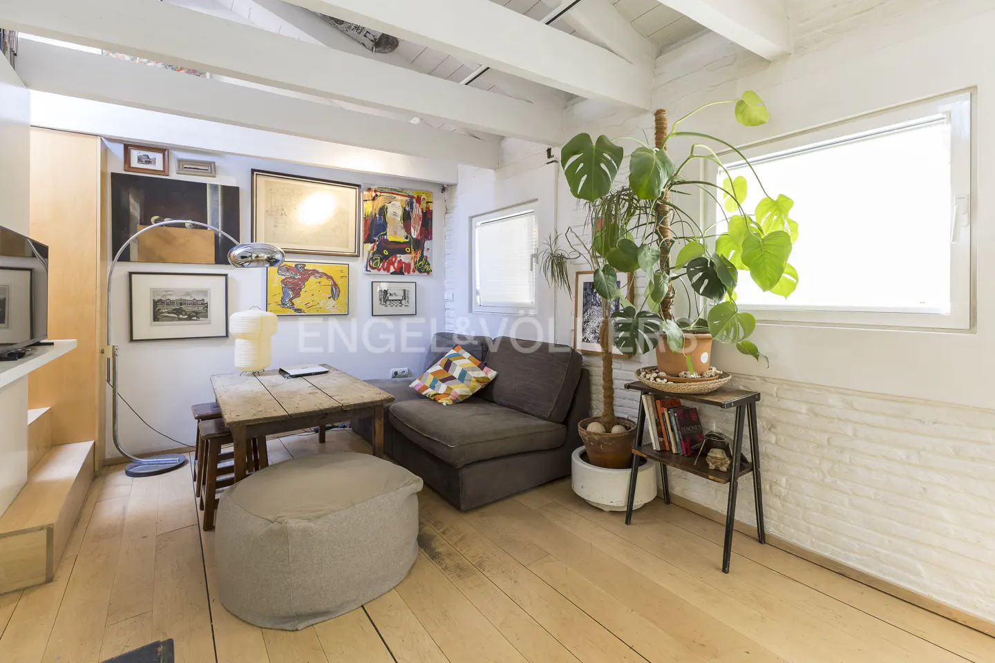 Cozy living room with wood floors, white walls, and exposed beams. A brown sofa, wooden table, and potted plants add warmth.