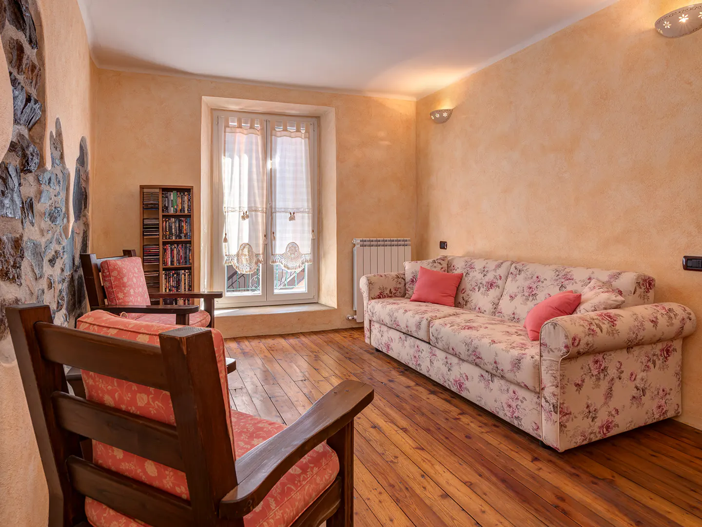 Cozy living room with floral sofa, wooden chairs, and a stone wall feature. Sunlight streams through the window.