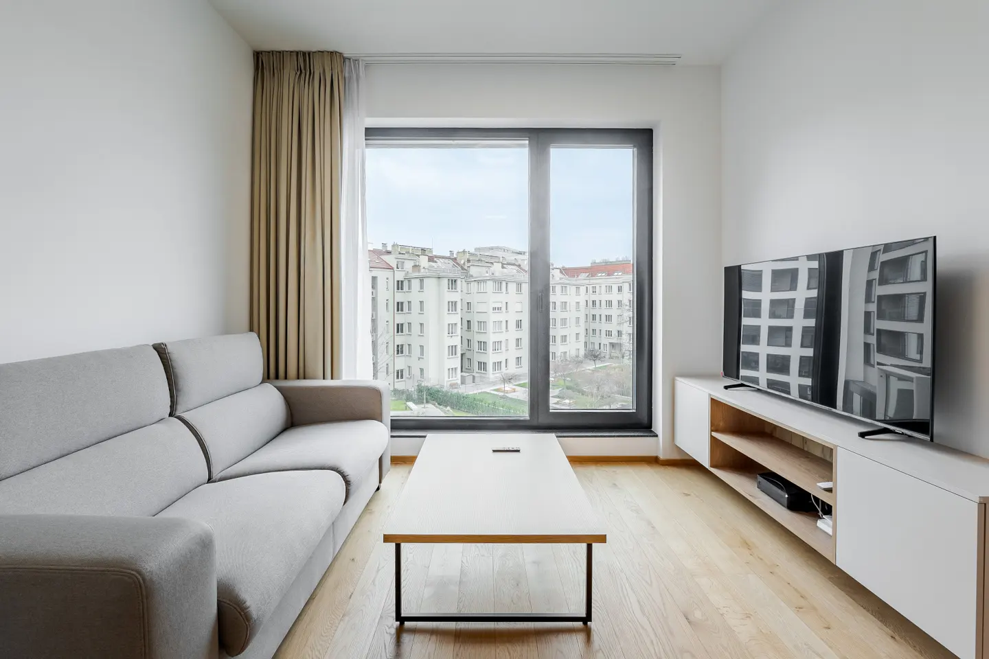 Modern living room with a grey sofa, white coffee table, media console, and a large window overlooking city buildings.