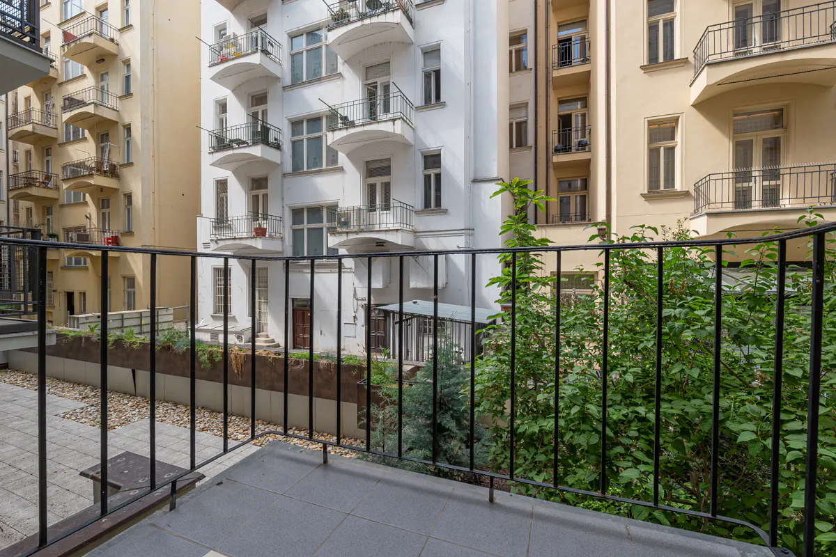 View from a balcony with black railings, overlooking a courtyard with plants and buildings in the background.