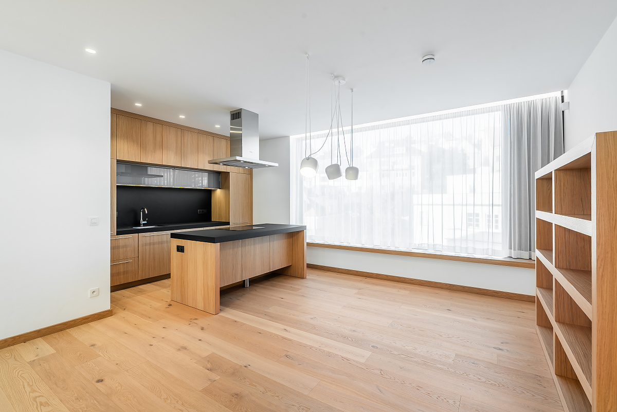 Modern kitchen with light wood cabinets, black countertops, a central island, and large windows with sheer white curtains.