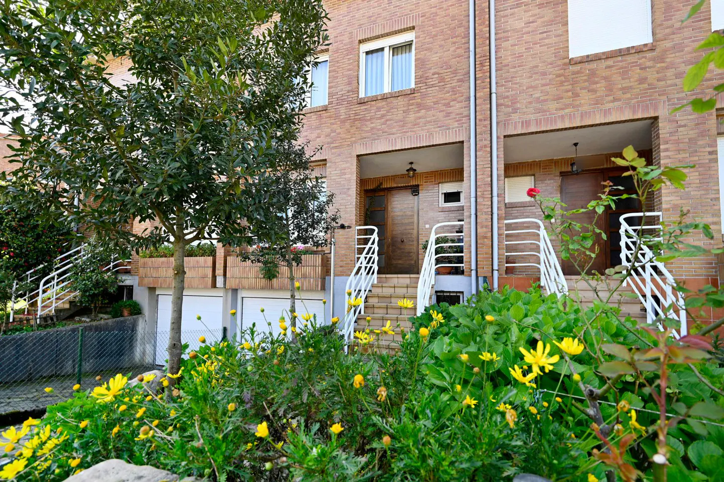 Brick townhouses with white railings lead to covered porches. Yellow flowers and greenery fill the front garden.
