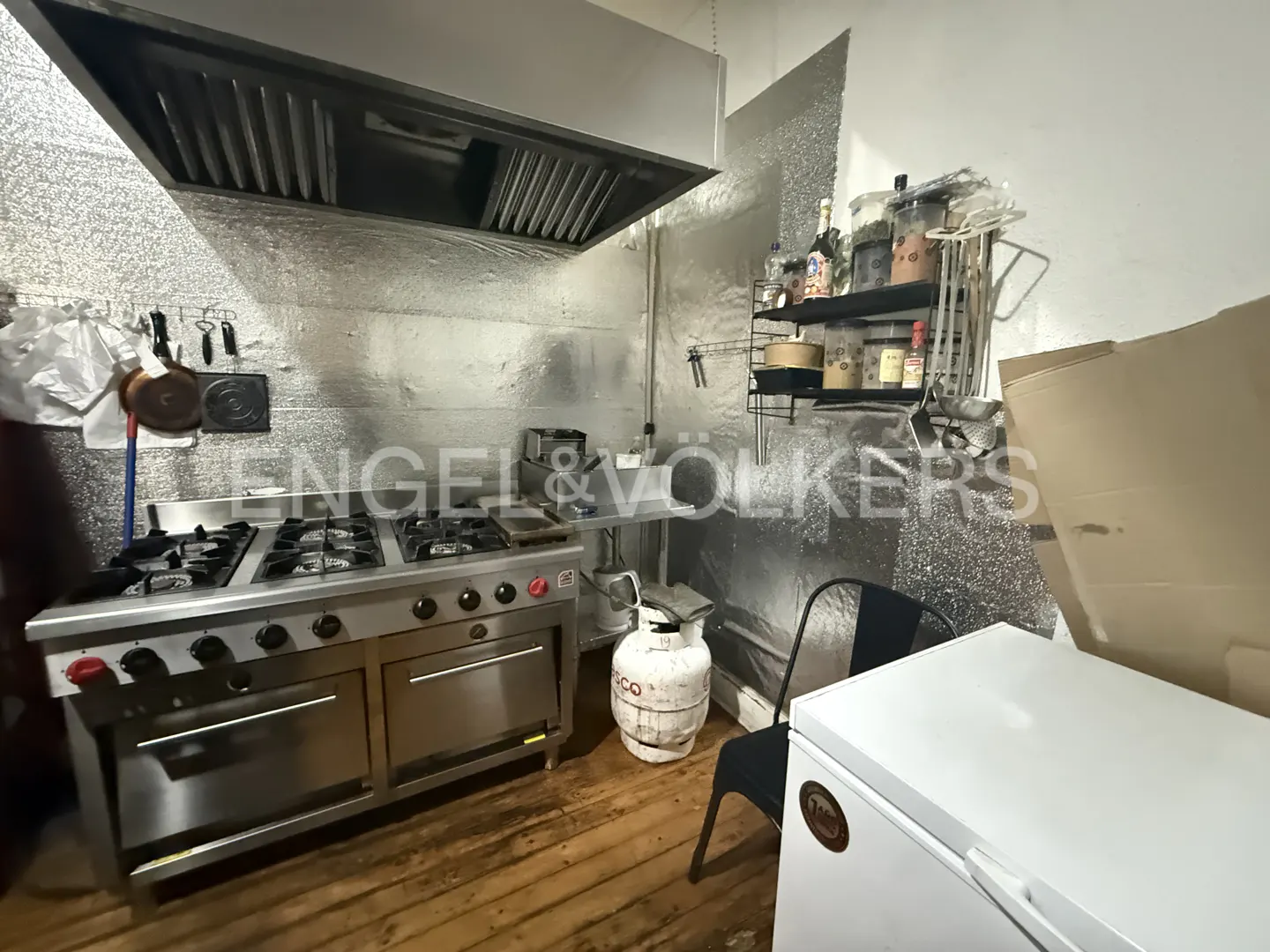 Commercial kitchen with a stainless steel gas range, large exhaust hood, propane tank, and a white chest freezer.