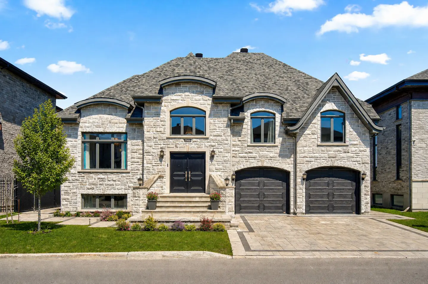 Two-story stone house with a gray roof, black double doors, and a two-car garage. Green lawn and blue sky.