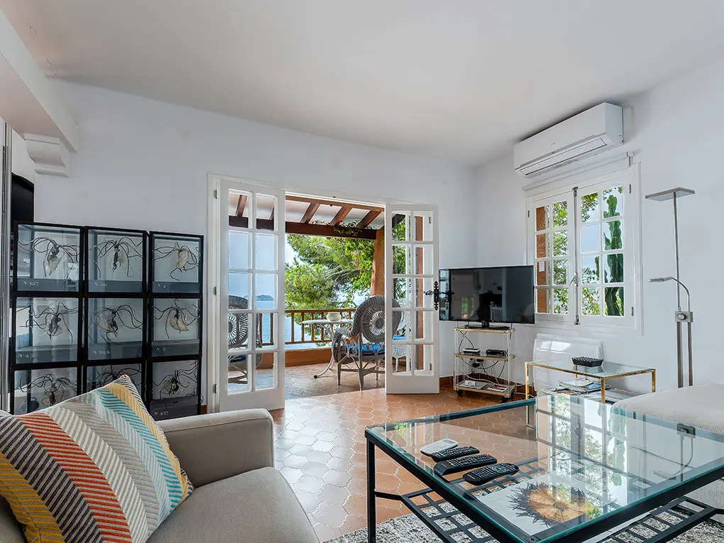 Bright living room with white walls, glass coffee table, TV, and open French doors to a balcony with ocean views.