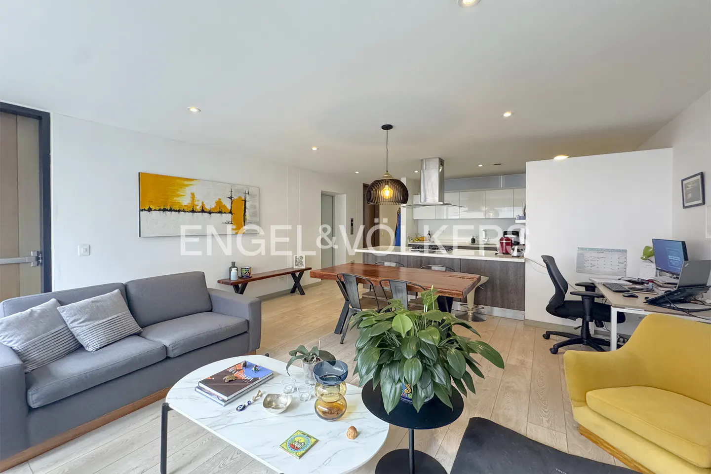 Bright, open-concept living space with gray sofa, wood table, kitchen, and desk area. A yellow armchair adds a pop of color.