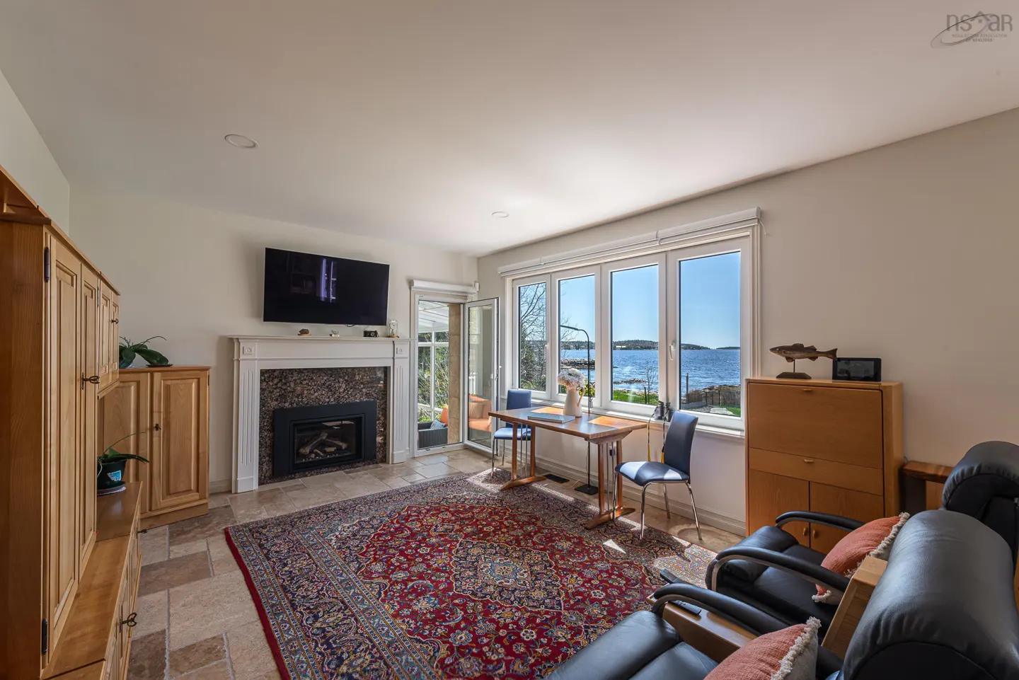 Living room with a fireplace, TV, and a view of the ocean through large windows. A red rug covers the tile floor.