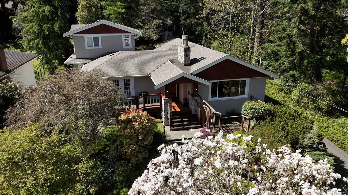 Aerial view of a gray two-story house with a stone chimney, surrounded by trees and flowering bushes.