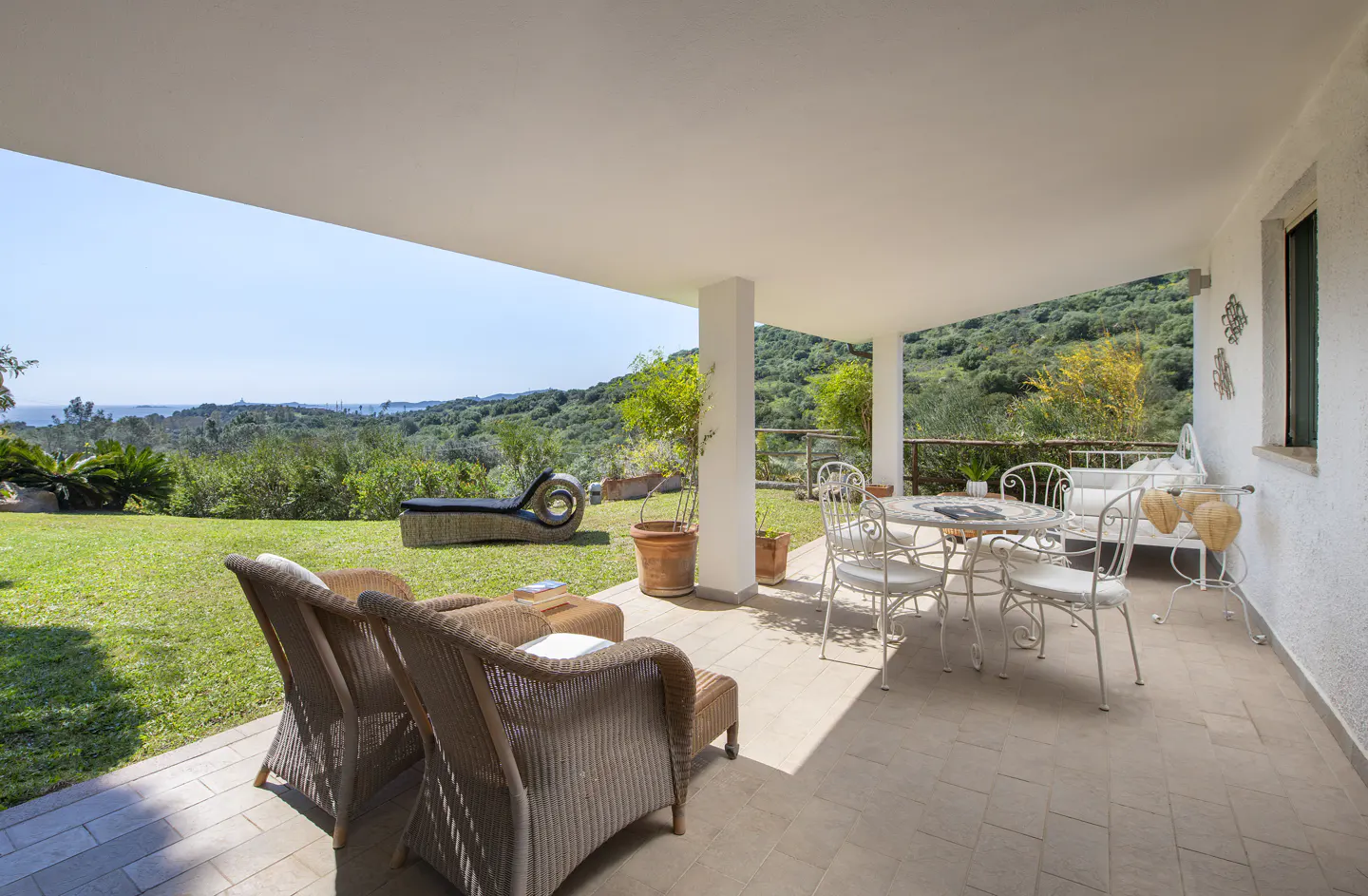 Covered patio with wicker furniture, a white table and chairs, and a chaise lounge on the lawn overlooking a green, hilly landscape.