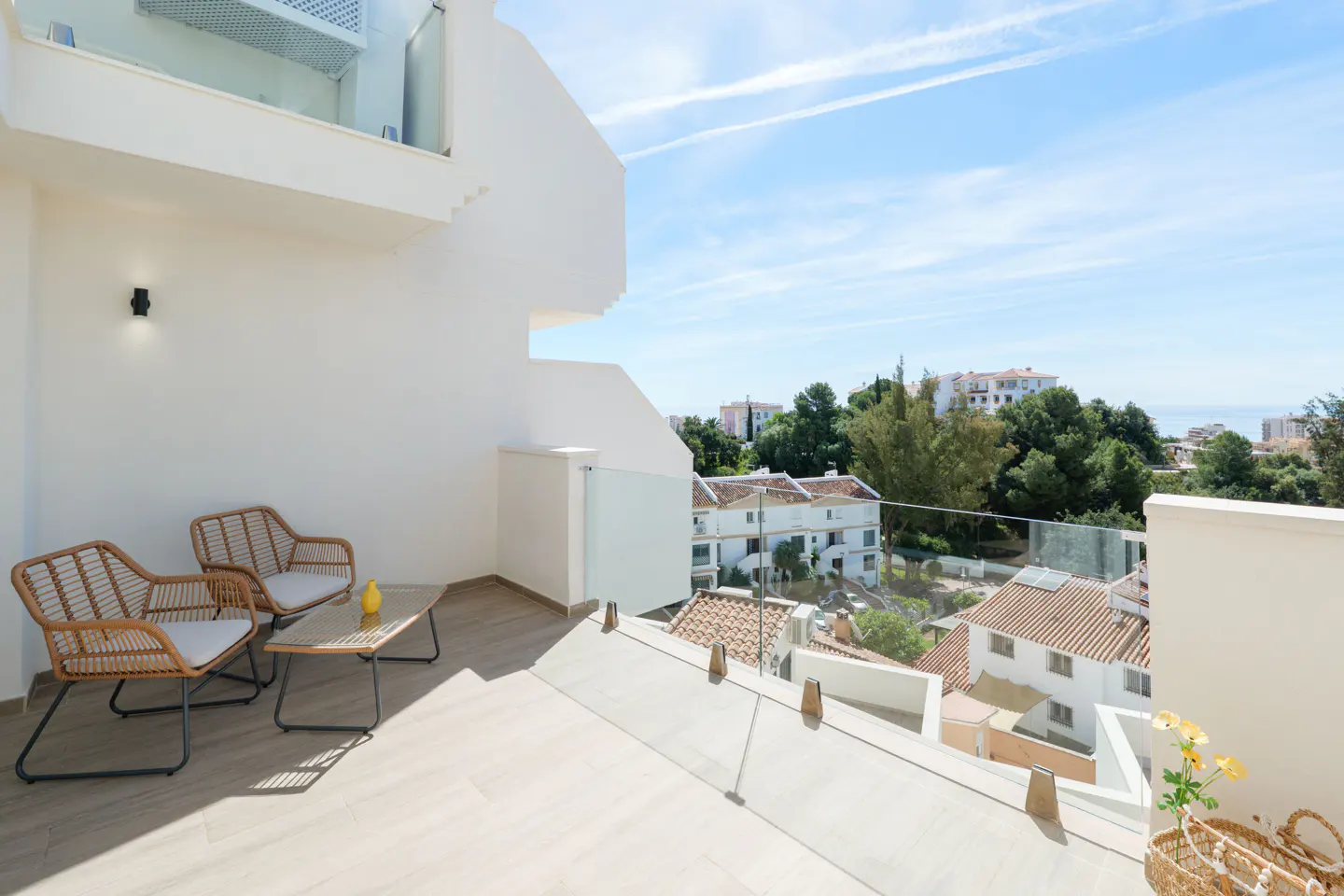 Balcony view with two wicker chairs, a table, and a basket of flowers overlooking a neighborhood with trees and a blue sky.