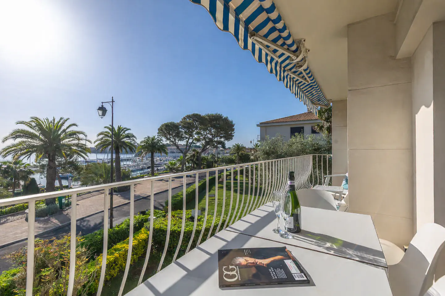 Balcony view with white table, champagne, and magazine. Palm trees and a marina are visible in the background under a blue sky.