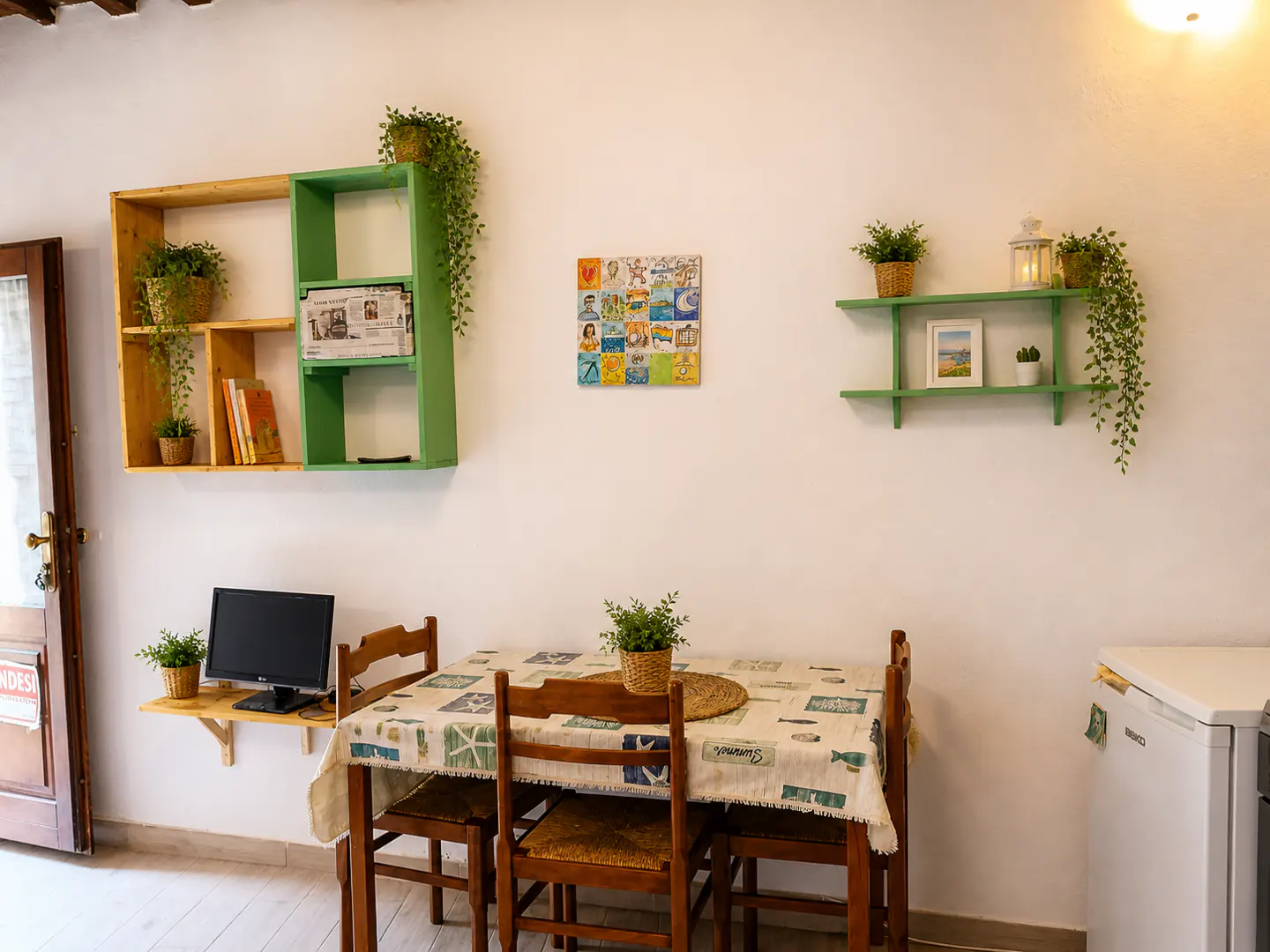 A dining area with a wooden table, chairs, and a patterned tablecloth. Shelves with plants and decor adorn the white walls. A computer sits on a small table.