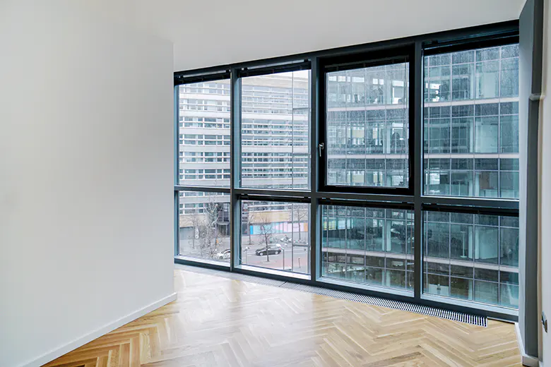 Empty room with herringbone wood floor and large windows. Outside, city buildings and street view. White walls.