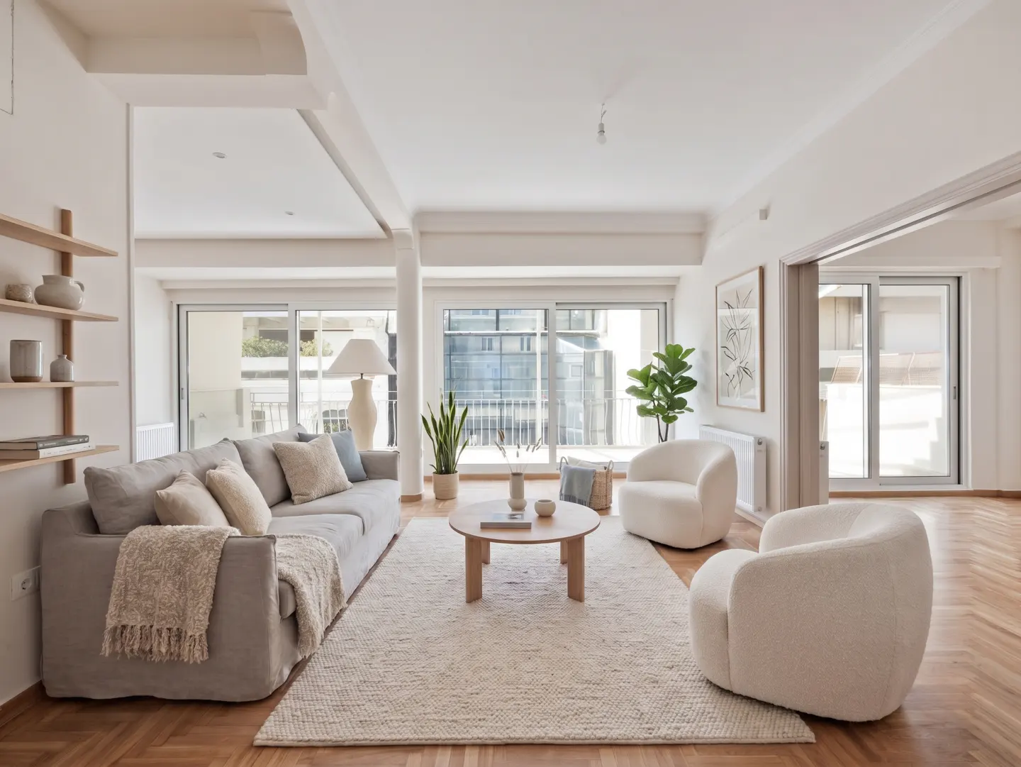Bright living room with a gray sofa, white armchairs, and a round coffee table on a cream rug. Large windows offer city views.