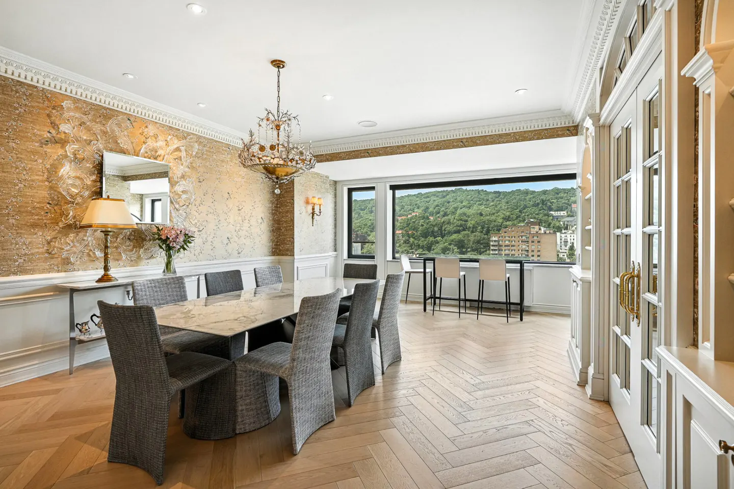 A dining room with a marble table, gray chairs, and a chandelier. A large window shows a view of trees and buildings.
