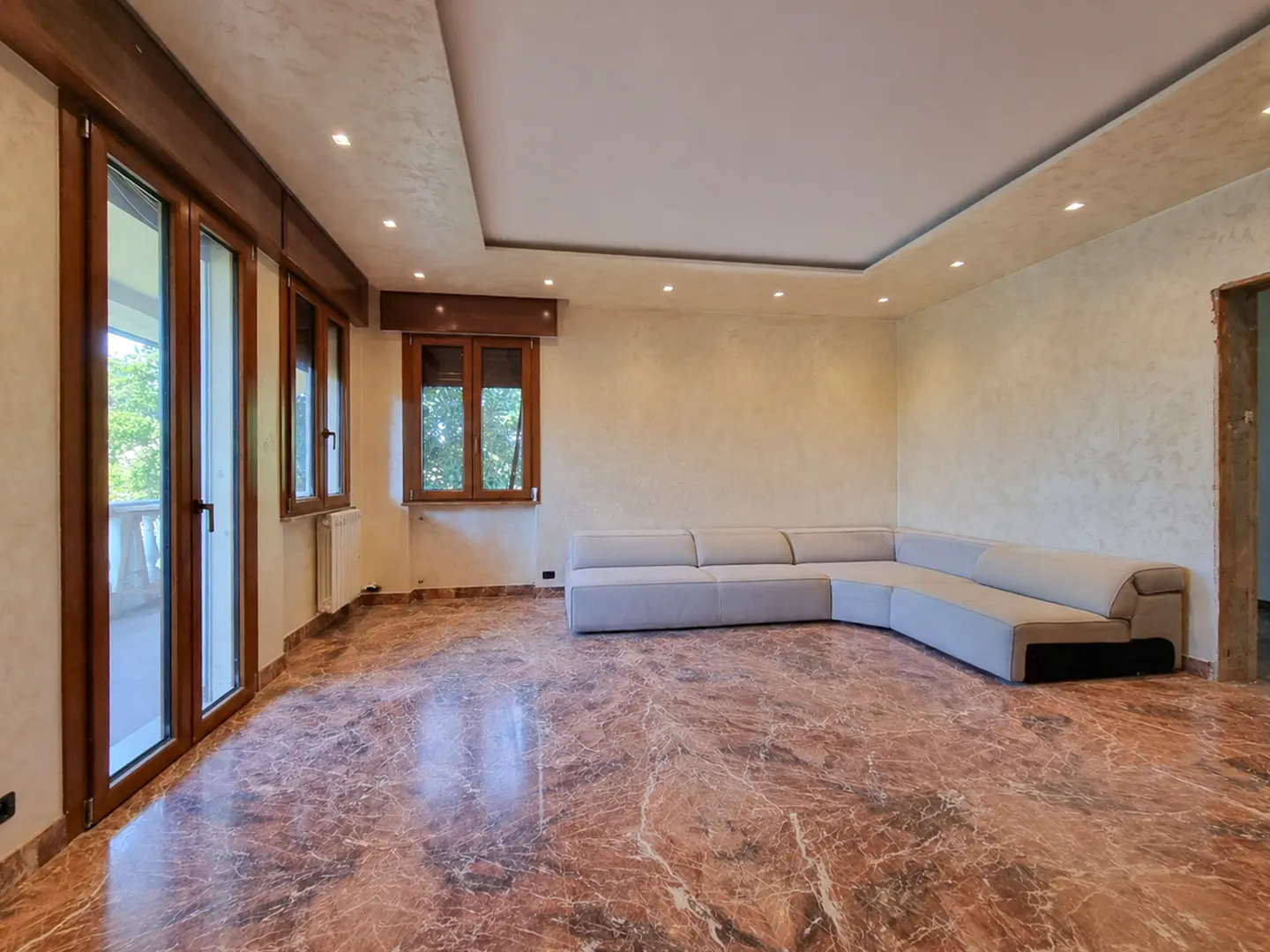 A spacious living room with marble floors, a light gray sectional sofa, and wood-framed windows.
