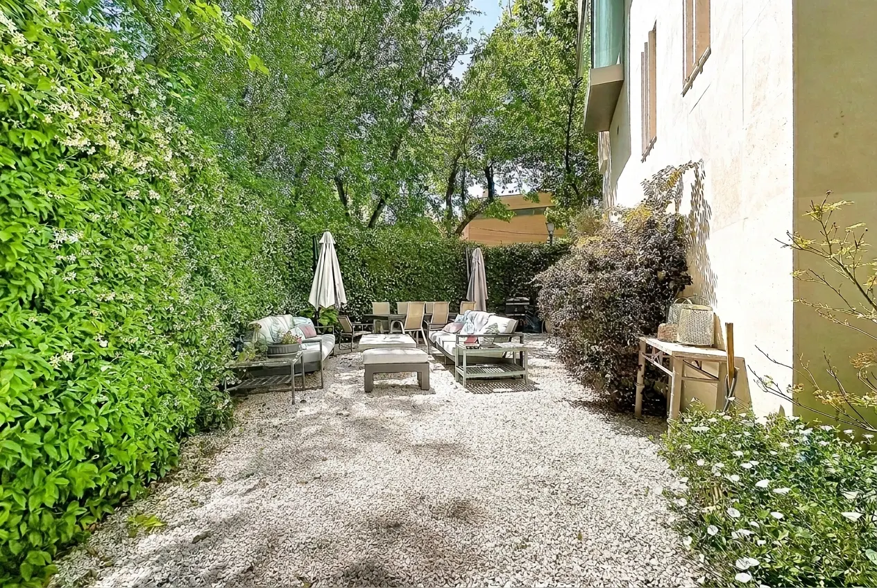 Outdoor patio with white gravel, surrounded by green foliage, lounge seating, and a dining table.