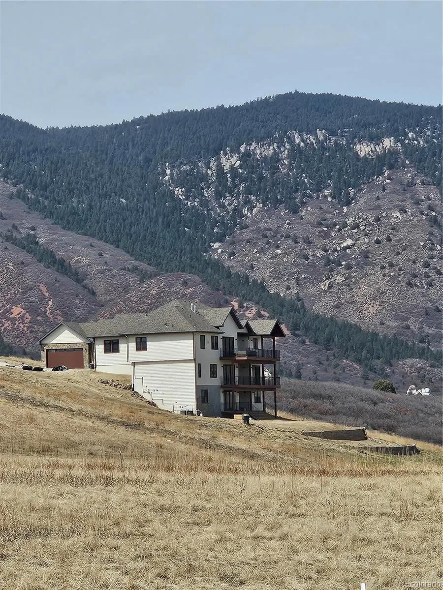 A three-story white house with balconies sits on a grassy hill with mountains in the background.