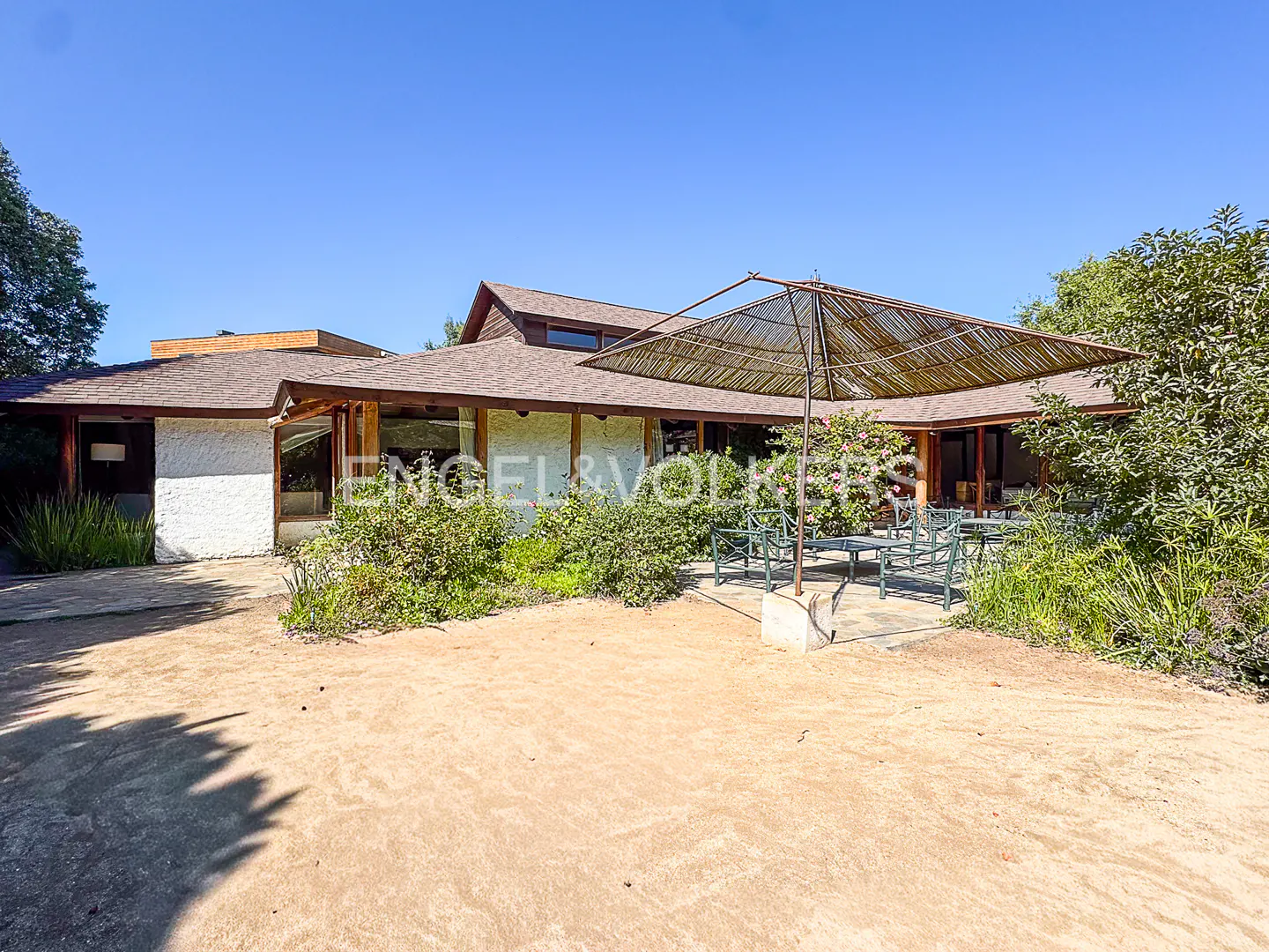 Exterior of a one-story house with a brown roof and white walls. A patio table with chairs sits under a large umbrella.