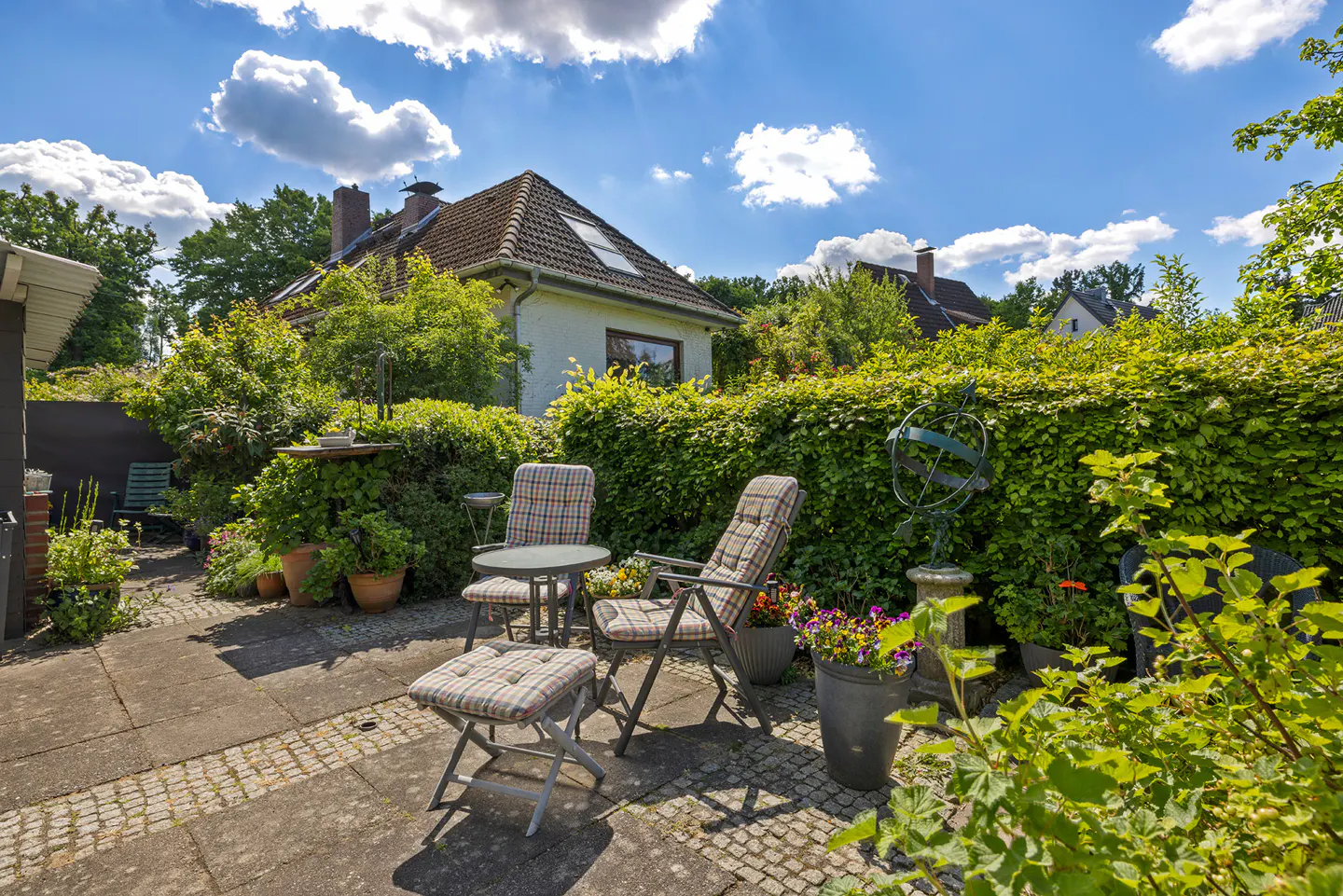 Outdoor patio with table, chairs, and footstool on stone pavers. Lush green hedges and trees surround the area. White house with brown roof in background.