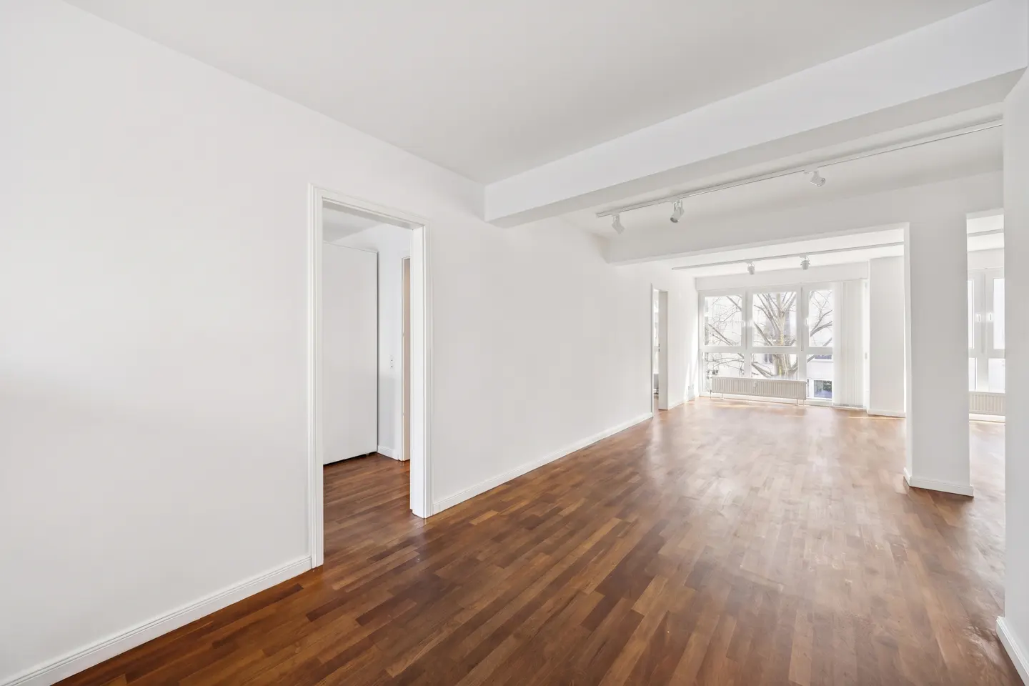 Bright, empty room with white walls, dark wood floors, and large windows. Track lighting on the ceiling. Doorway to the left.