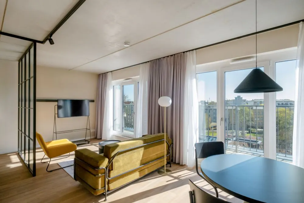 Bright apartment interior with a green sofa, yellow chair, black table, and large windows overlooking a city.