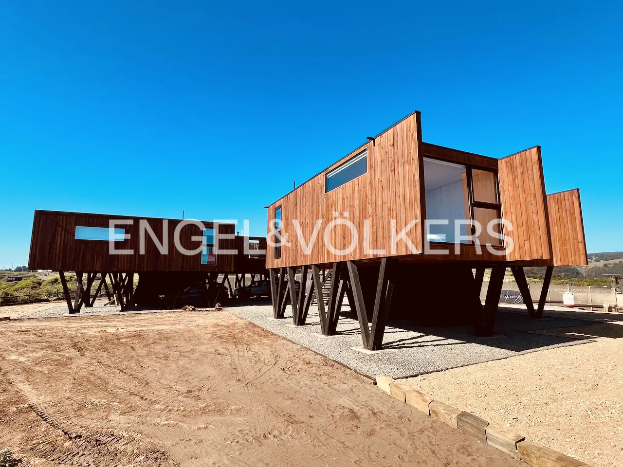 Exterior view of a modern, elevated wooden house with black supports under a clear blue sky.