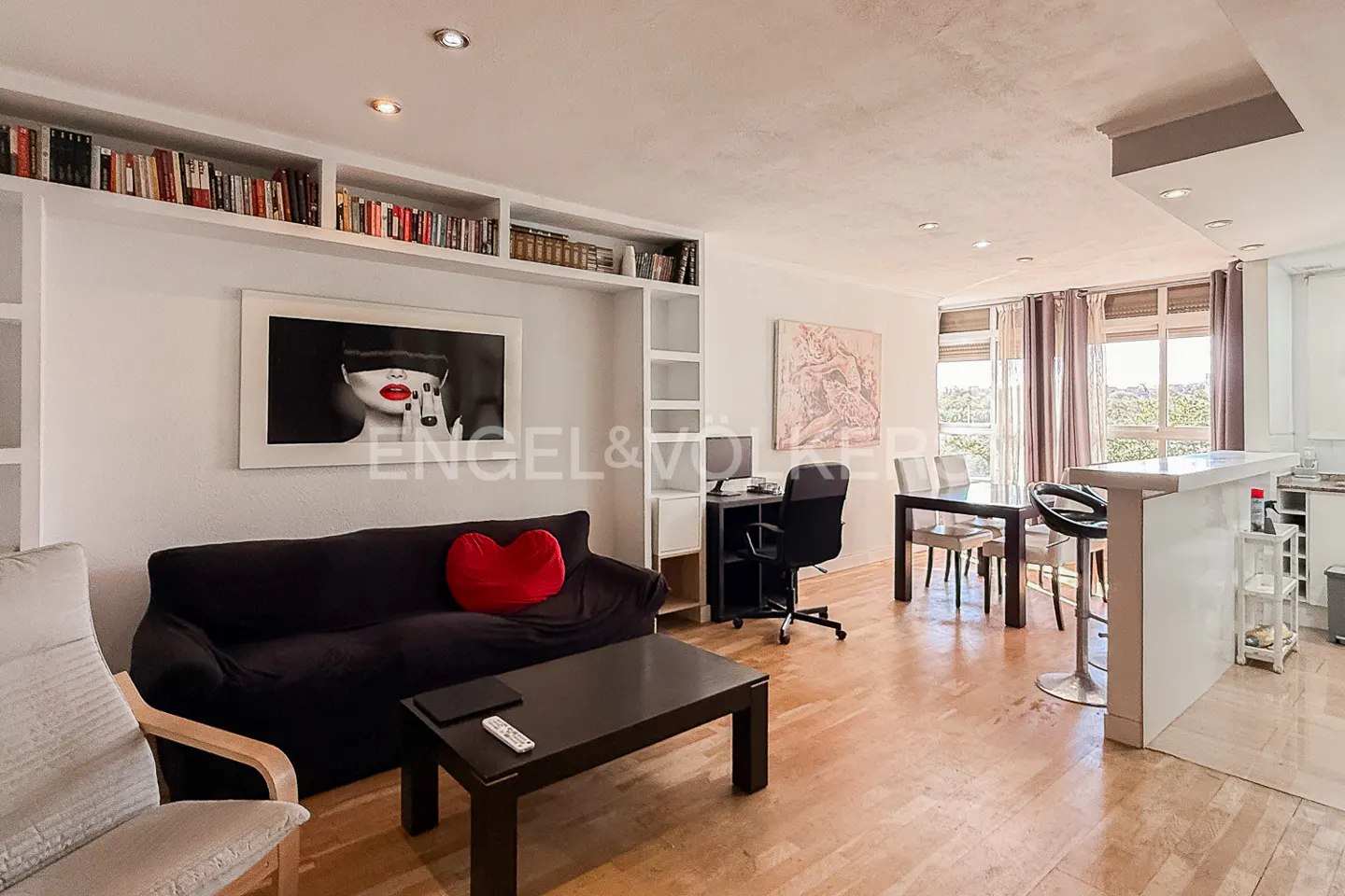 Bright living room with hardwood floors, black sofa with red heart pillow, and a black coffee table. Bookshelves line the wall above the sofa.