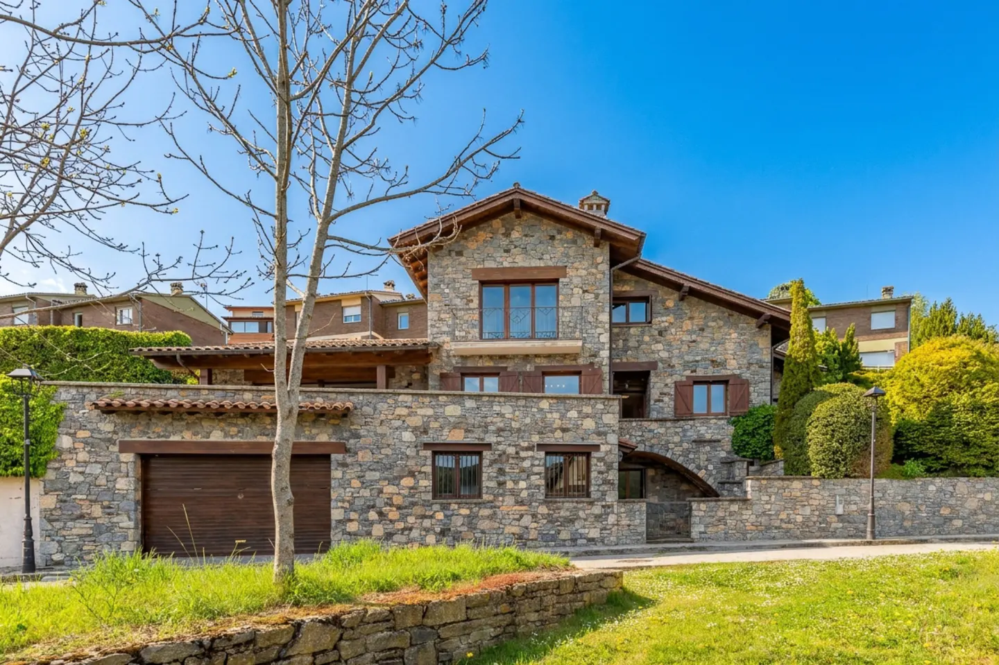 Stone house with brown shutters and garage door, green lawn, and blue sky.