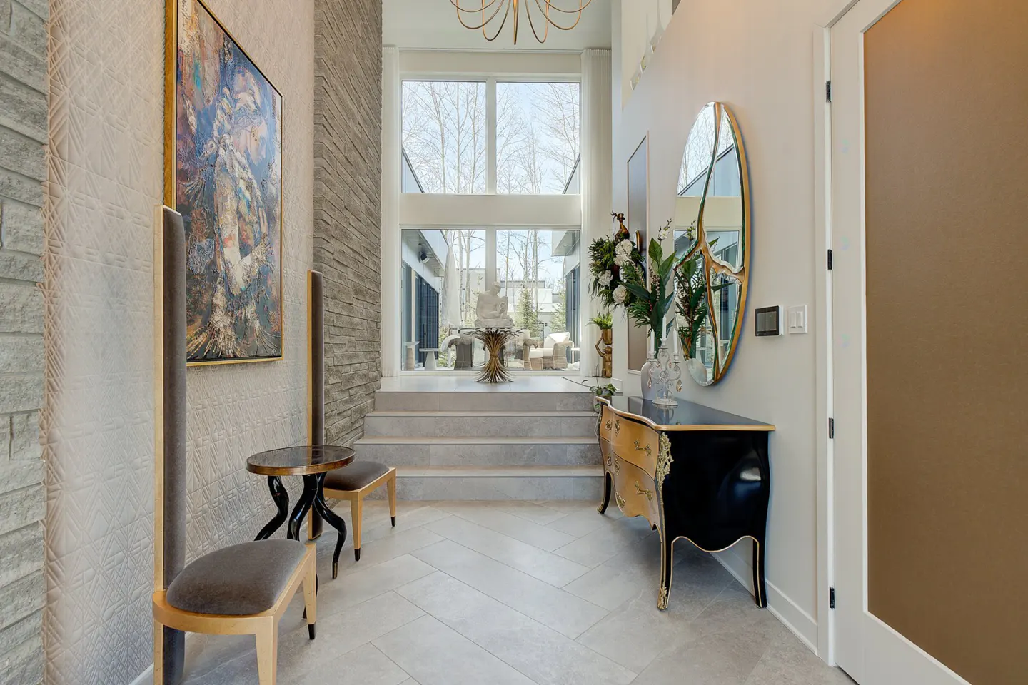Foyer with herringbone tile floor, stone accent wall, and large windows. A black and gold console table sits beneath an oval mirror. Artwork adorns the wall.