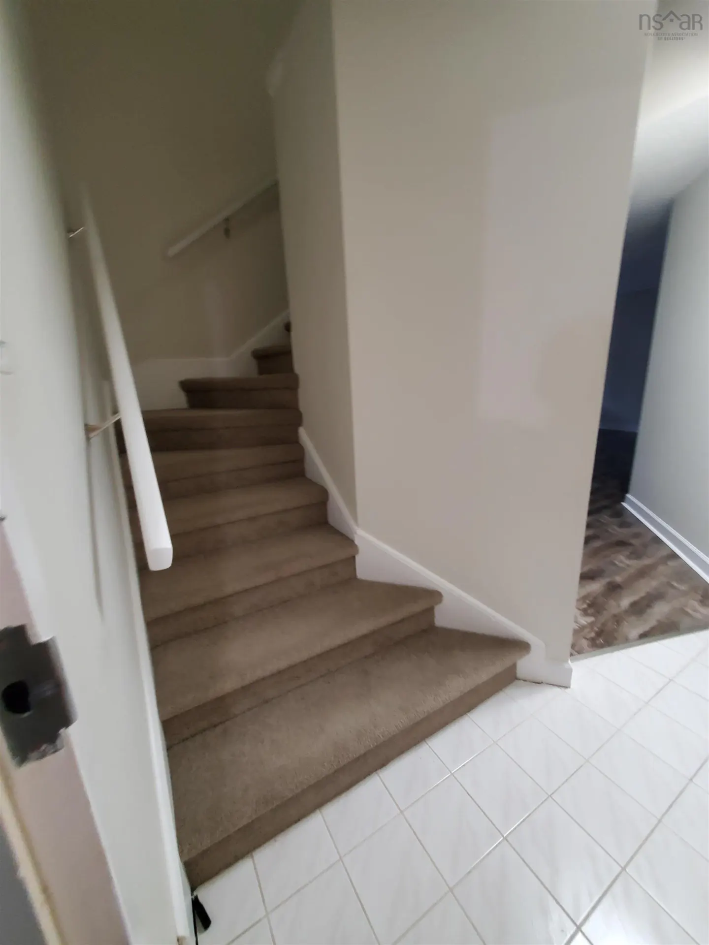 Interior view of a home featuring carpeted stairs with white walls and trim, leading to a hallway with tile flooring.