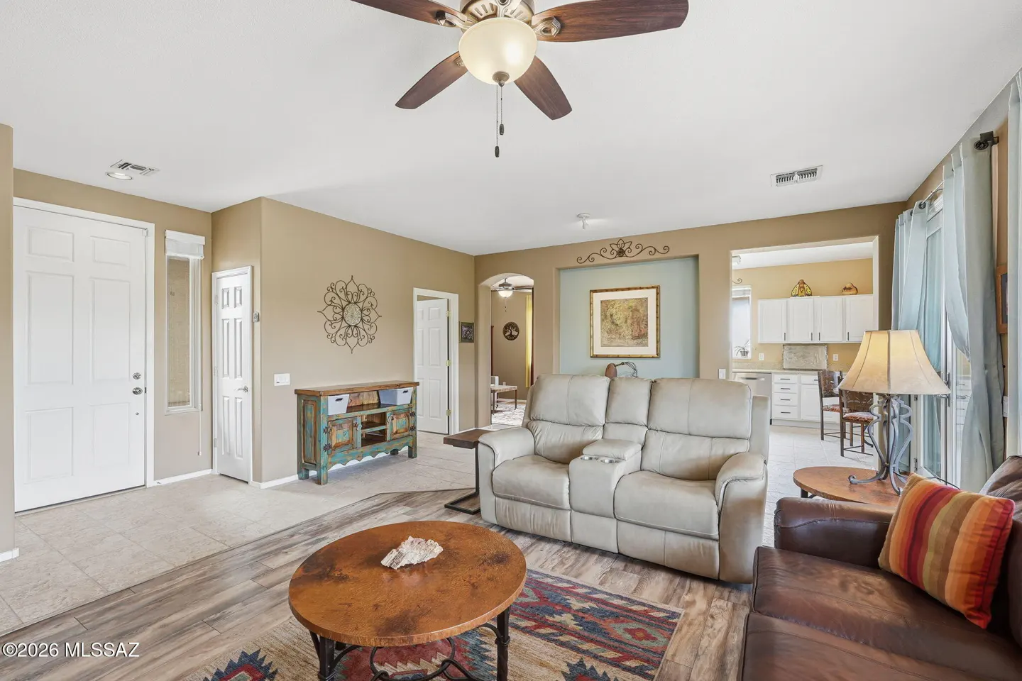 Living room with beige walls, a ceiling fan, and a tan leather loveseat. A round wooden table sits on a patterned rug. White kitchen in the background.