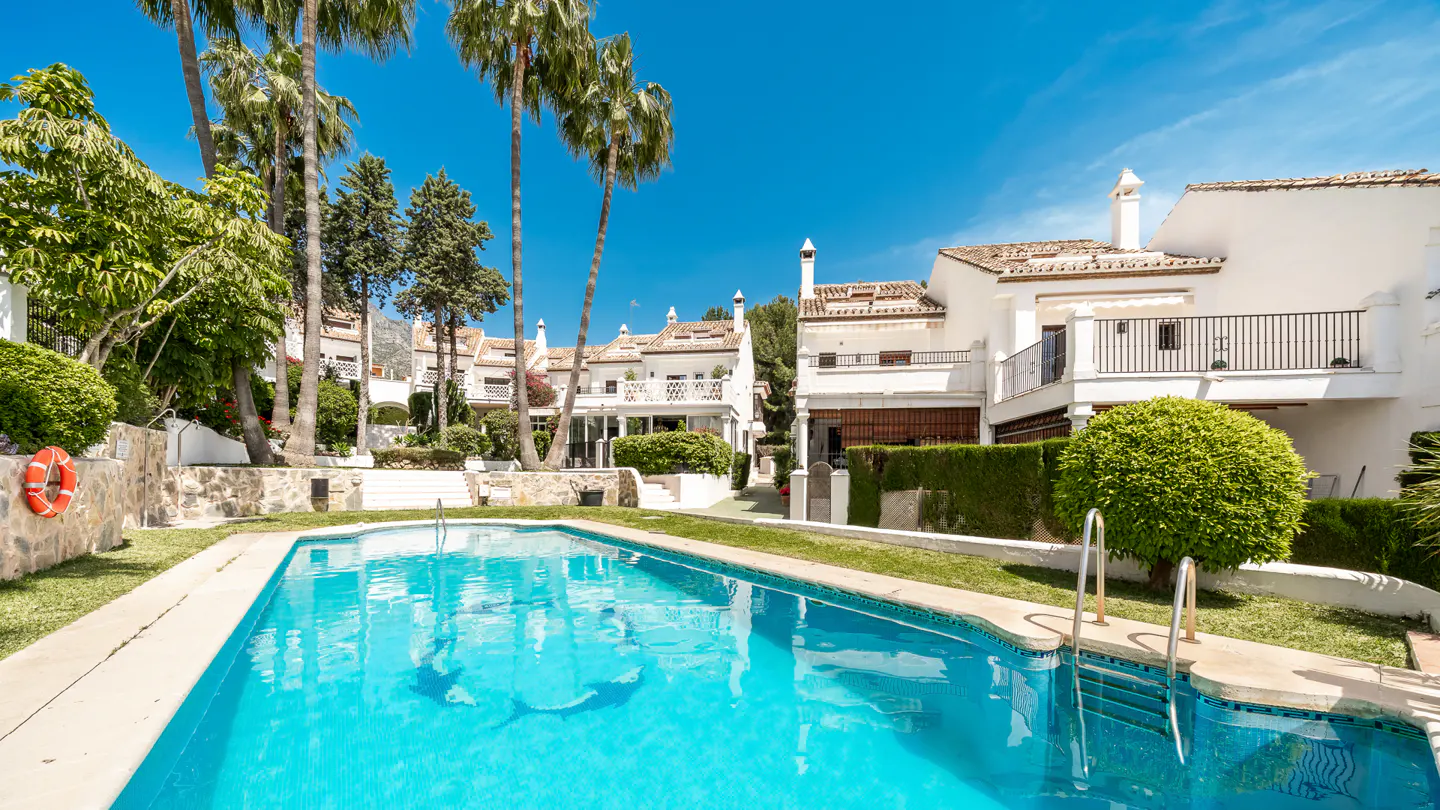 A turquoise pool is surrounded by white buildings, green grass, and palm trees under a blue sky.