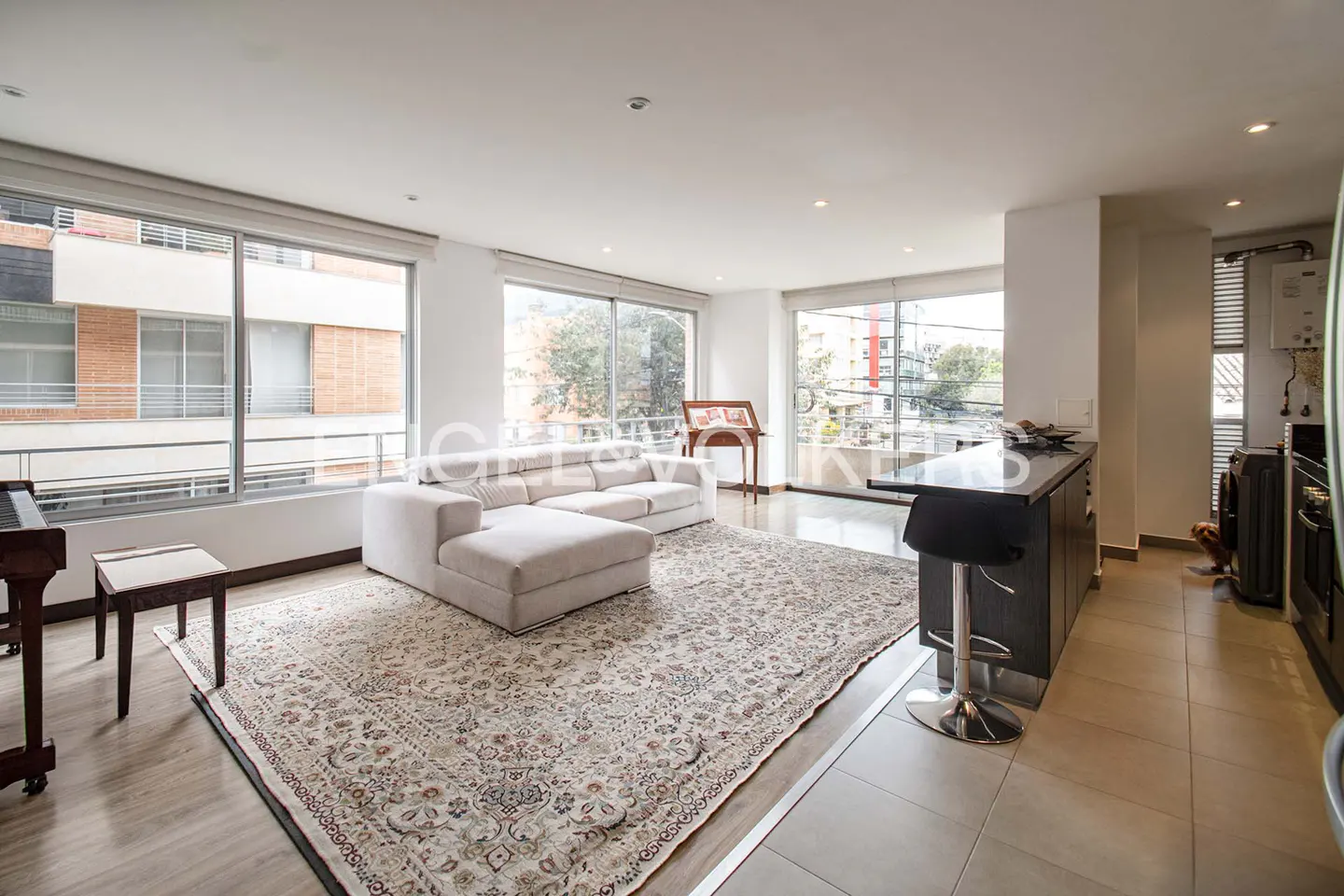 Bright, open-concept living space with a white sectional sofa on a patterned rug. Large windows offer city views. A kitchen island with bar stools is visible.