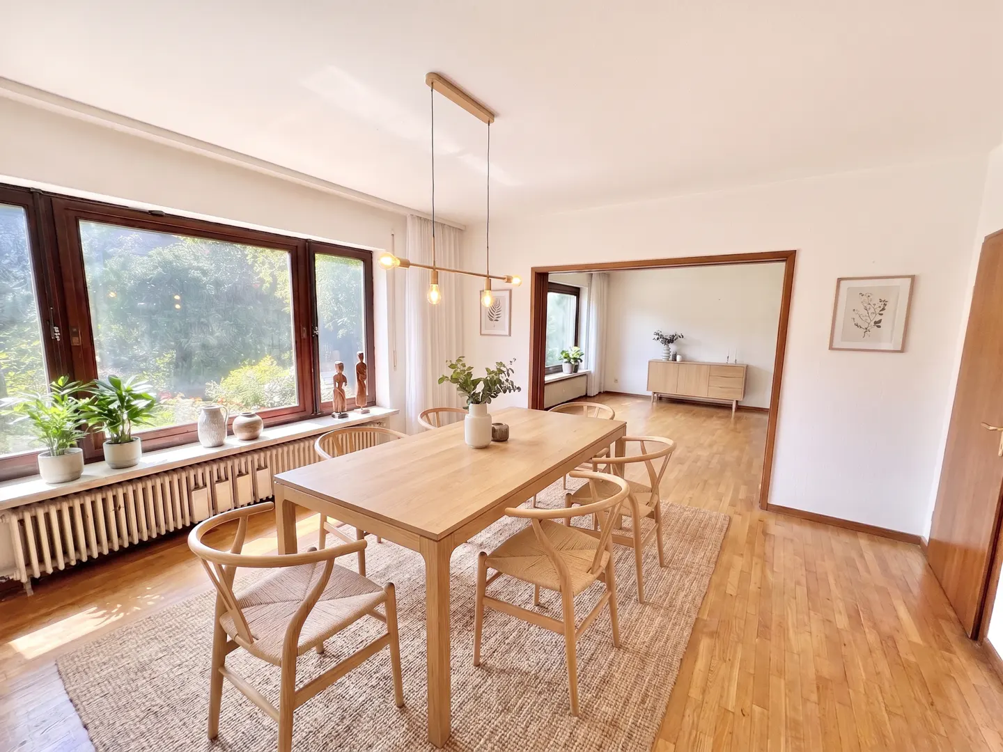 Bright dining room with a wooden table, six chairs, and a large window overlooking greenery. A modern light fixture hangs above the table.