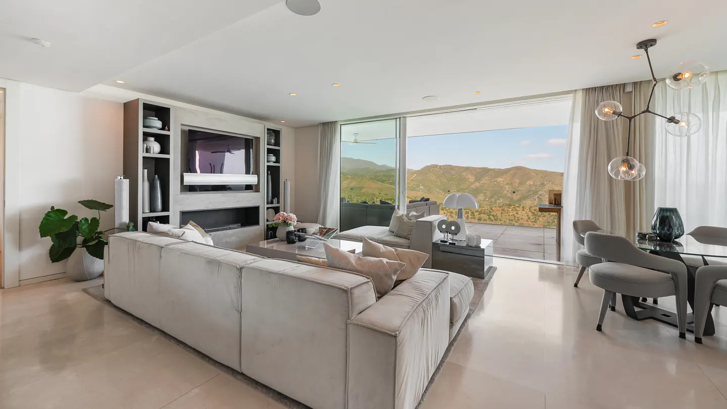 Bright, modern living room with a large gray sofa, TV, and mountain views through sliding glass doors.