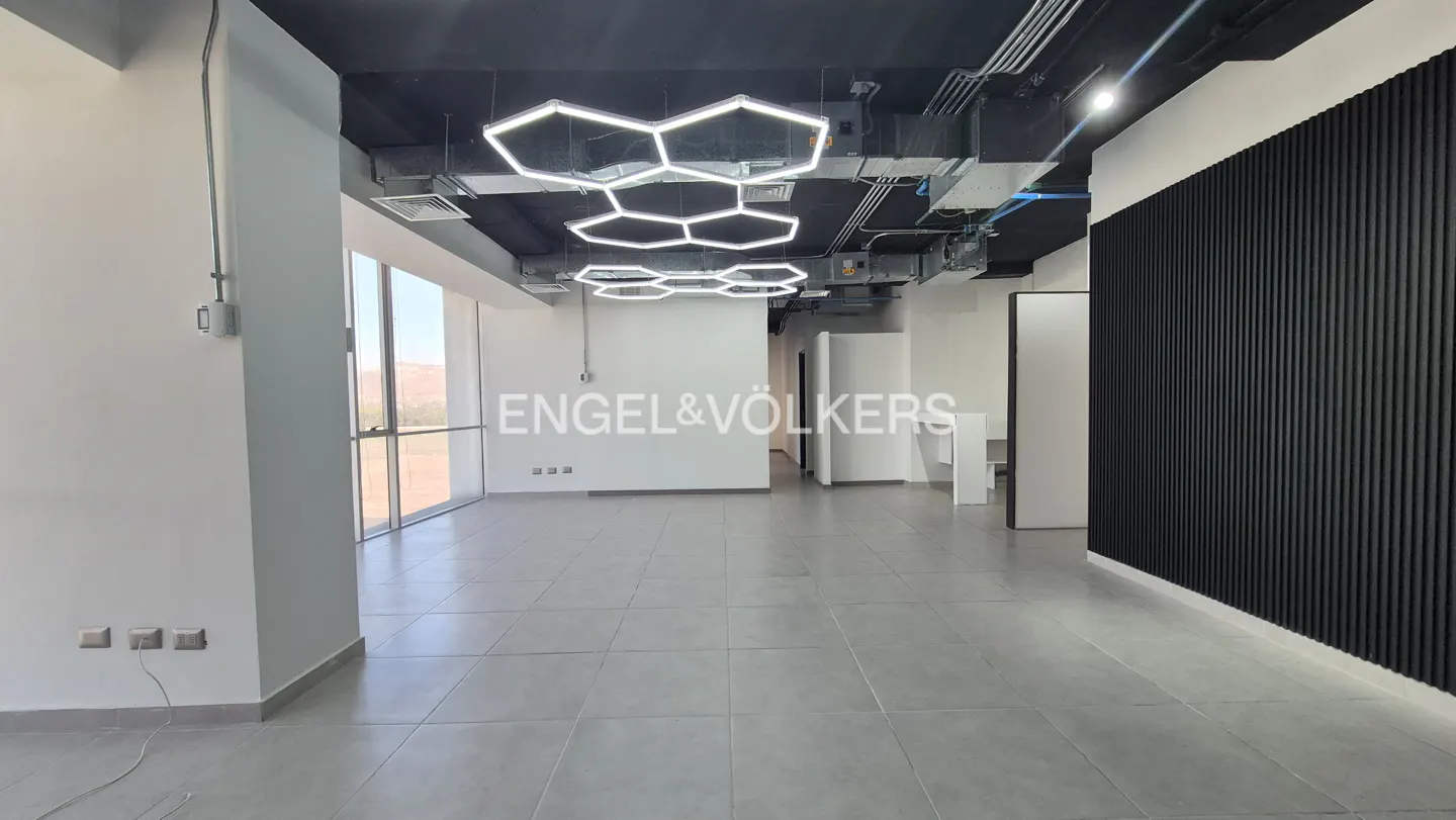 An empty office space with gray tile floors, white walls, and honeycomb-shaped lights hanging from a black ceiling.