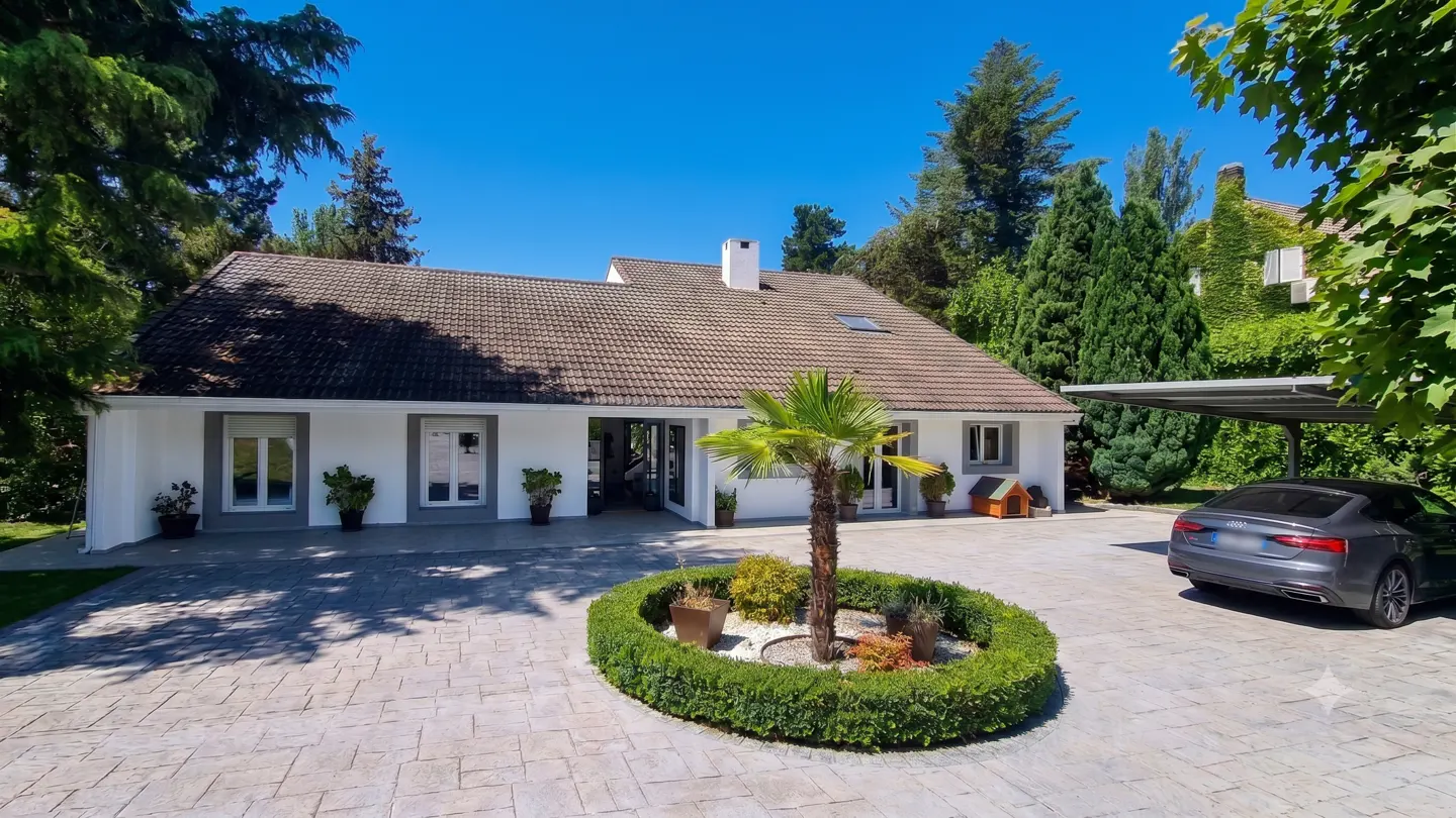 White single-story house with a brown tiled roof, paved driveway, central palm tree, and a parked grey sedan.