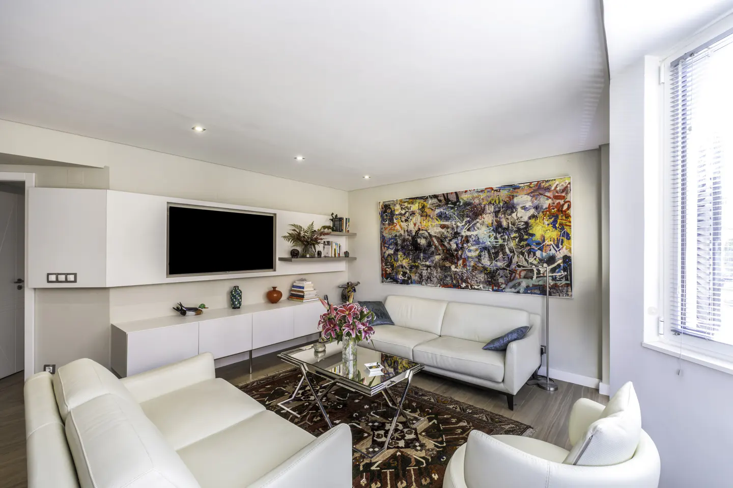 Bright living room with white sofas, a glass coffee table with pink flowers, and a colorful abstract painting on the wall. A large TV is mounted above white cabinets.