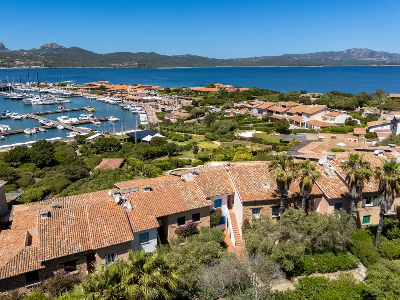 Aerial view of a coastal town with terracotta roofs, a marina filled with boats, and a blue sea with mountains in the background.