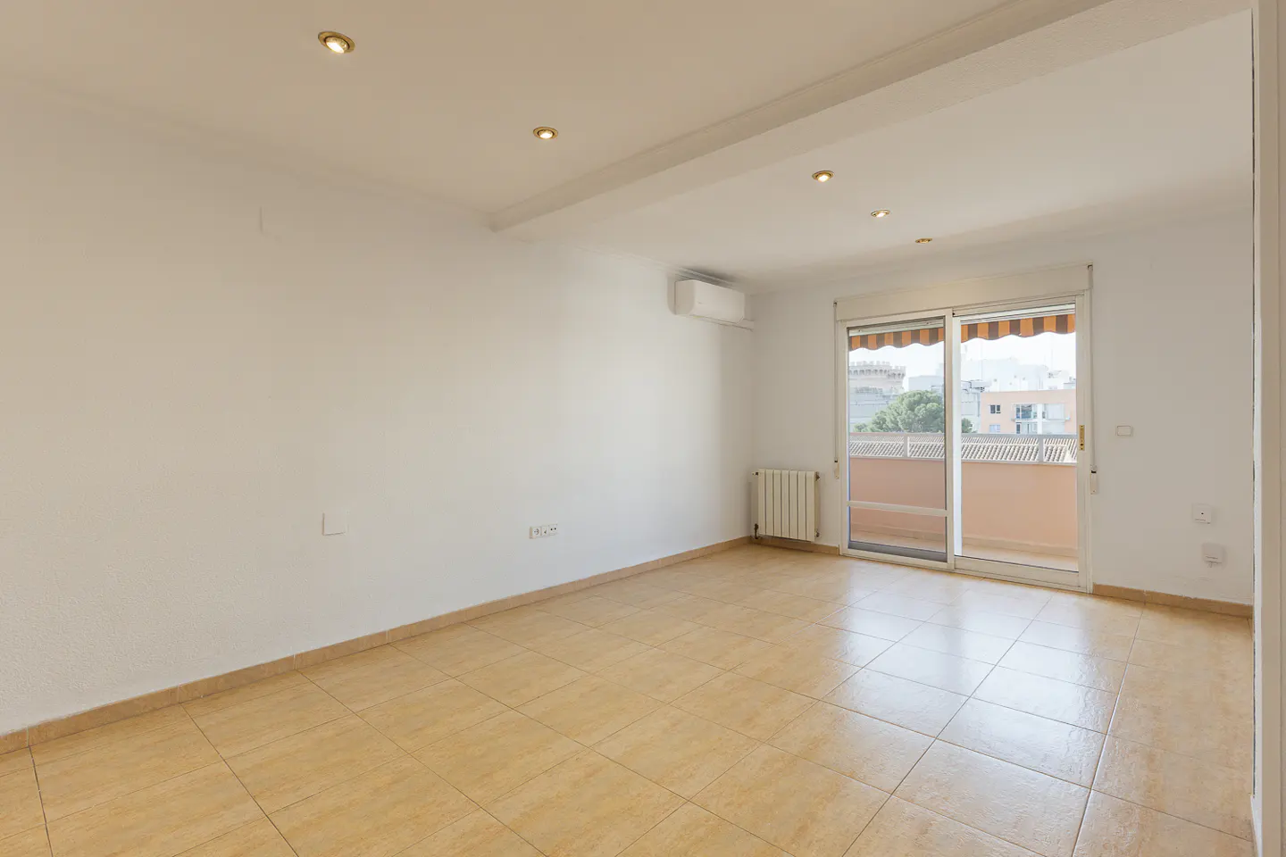 Bright, empty room with beige tile floor, white walls, and sliding glass doors to a balcony. Recessed lighting and an air conditioner are visible.