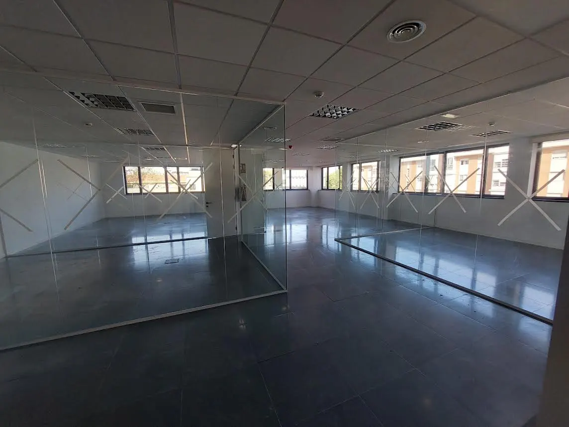 Empty office space with glass walls, gray tile floor, and white ceiling. Windows line the back wall, letting in natural light.