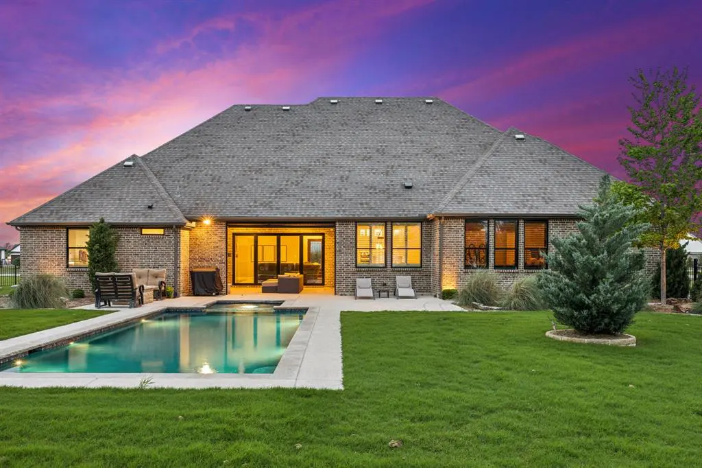 Backyard view of a brick house with a pool, green lawn, and a purple and pink sky.
