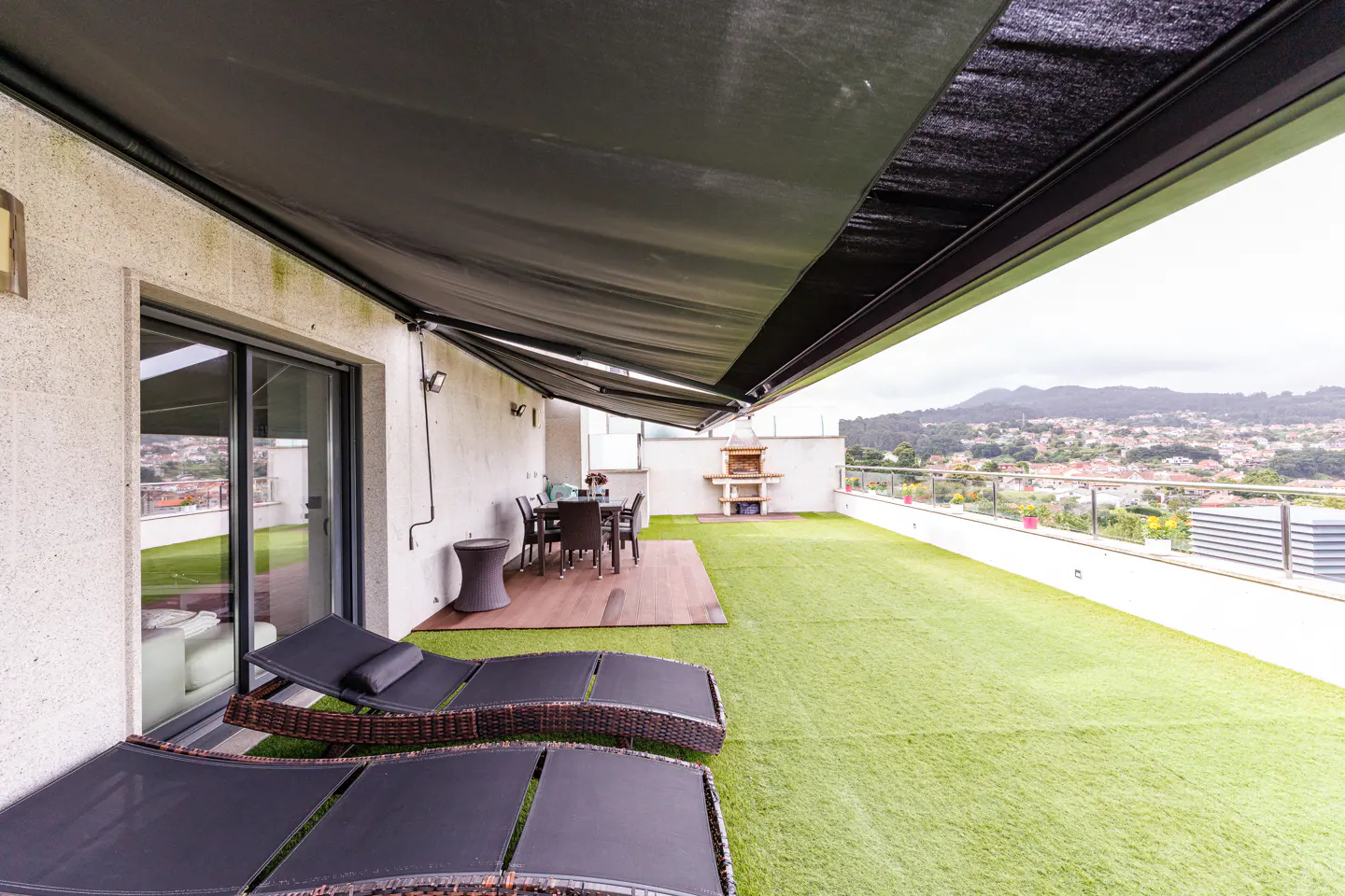 Spacious rooftop terrace with artificial grass, two lounge chairs, a dining set, a brick barbecue, and a retractable awning.