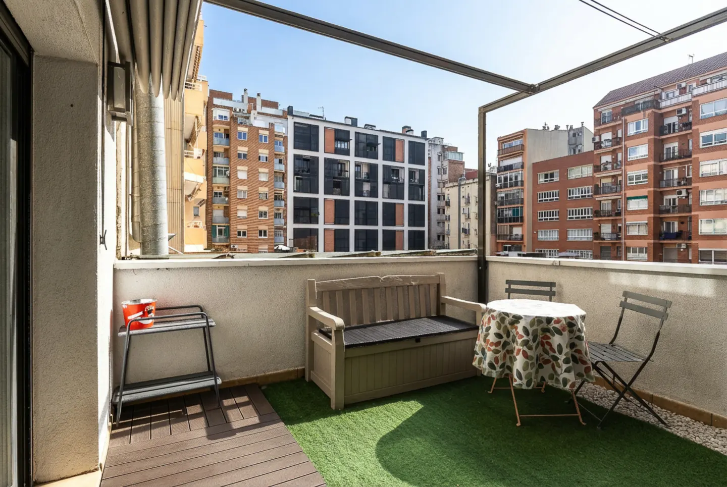 A balcony with a bench, table, and chairs overlooks city buildings under a blue sky.