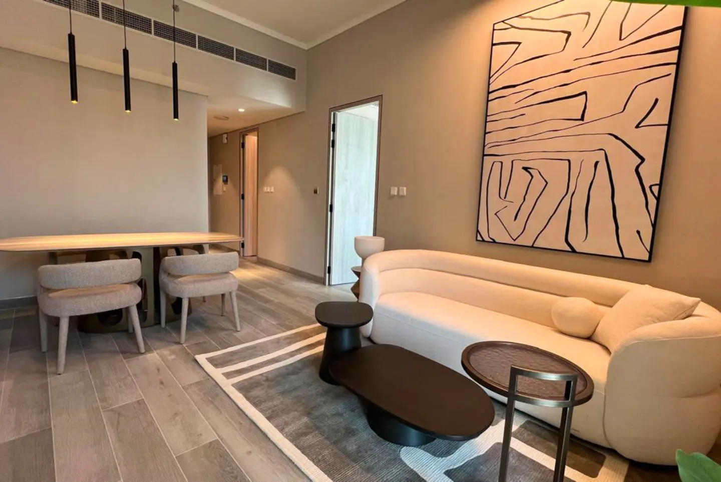 A modern living room with a beige sofa, black coffee tables, and a large abstract painting on the wall. A dining table with two chairs is visible in the background.