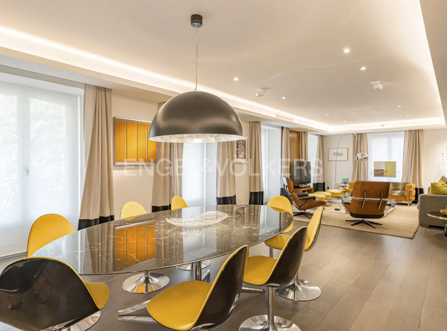 A modern dining room with a black marble table, yellow chairs, and a large dome pendant light. The room opens to a living area with brown leather chairs.
